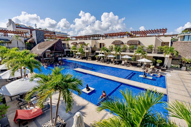 Long blue shared pool seen from above, with palm trees, lounge chairs, umbrellas and the building around it