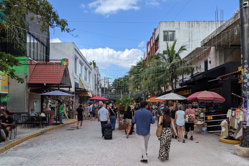 People walking along the pedestrian 5th Avenue past shops, cafe tables and red umbrellas, with palm trees and string lights overhead