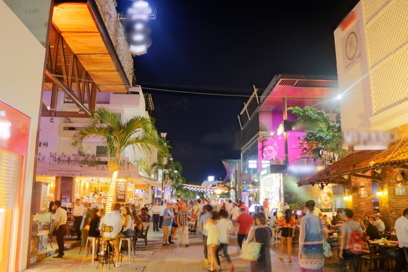 Crowds walking 5th Avenue at night past lit restaurants and bars with outdoor tables, a palm tree and bright signs