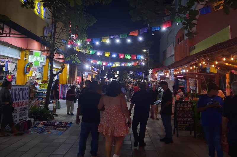 People strolling a paved stretch of 5th Avenue at night under strings of lights and paper banners, with shops open on both sides