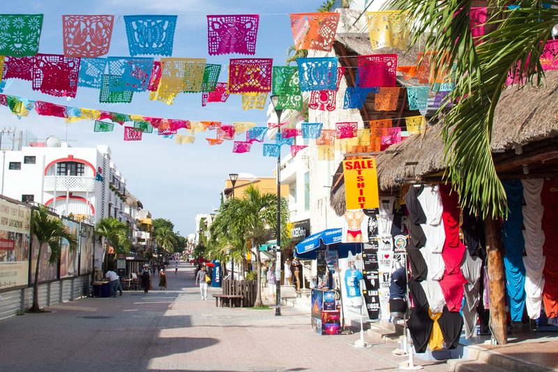 Rows of colorful cut-paper banners strung across 5th Avenue, with T-shirt stalls under a thatched roof and palm trees along the street