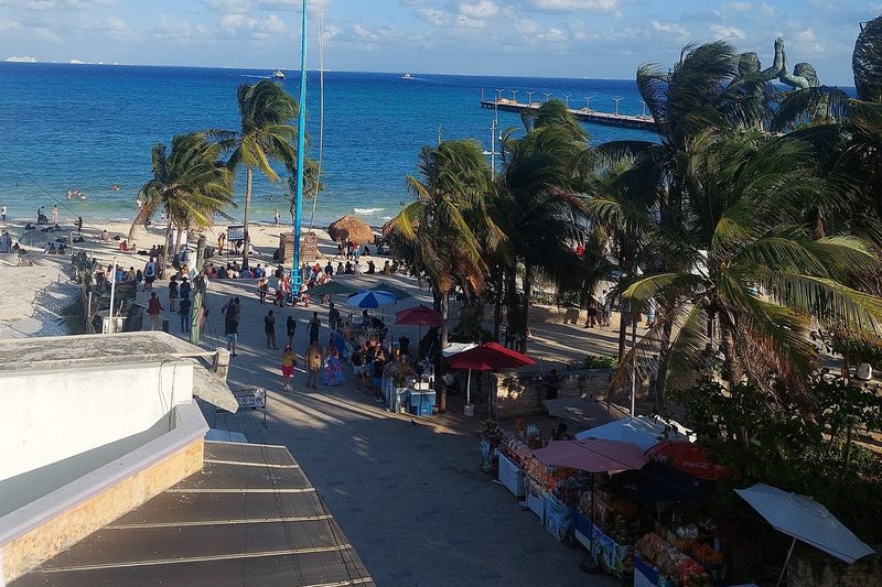 View from above of the beach at Parque Fundadores with palm trees, market stalls, a tall pole, people on the sand and a long pier into the turquoise sea