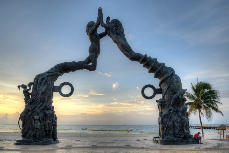 Bronze arch sculpture of two figures reaching toward each other on the waterfront, with the sea, a pier and a palm tree under an early morning sky