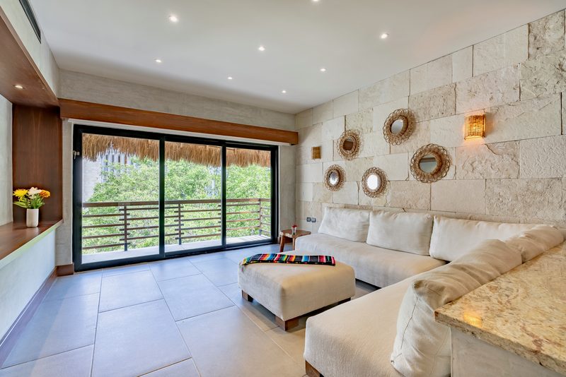 Living room with a cream sectional, an ottoman with a striped blanket, a stone wall with round woven mirrors and sliding glass doors to a balcony