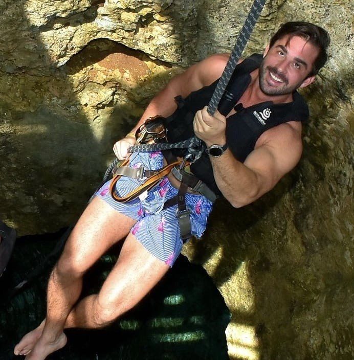 Isaac sonriendo mientras hace rappel por una cuerda hacia un cenote, con arnés y chaleco negro, entre paredes de roca