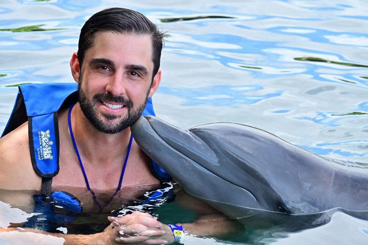 Isaac sonriendo en el agua de Xel-Há con un chaleco salvavidas azul, con un delfín apoyando la cabeza en su hombro