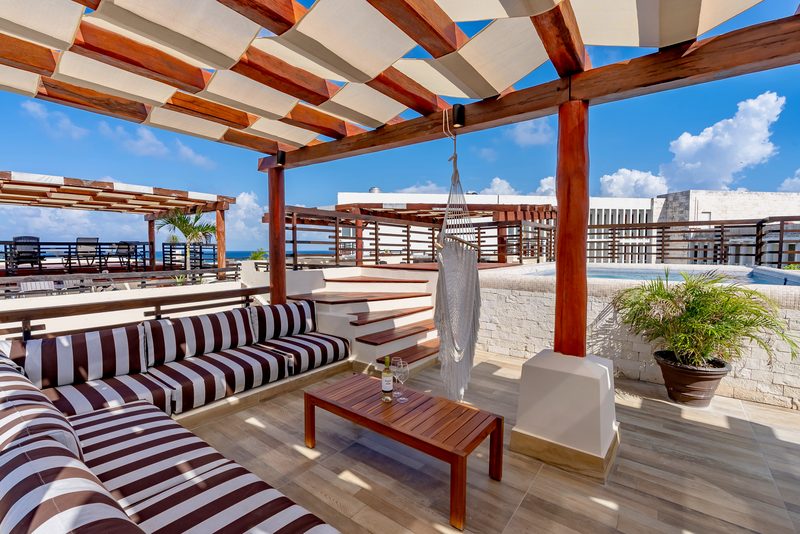 Brown and white striped lounge sofas around a wood coffee table under the rooftop pergola, with a hanging hammock chair and steps behind