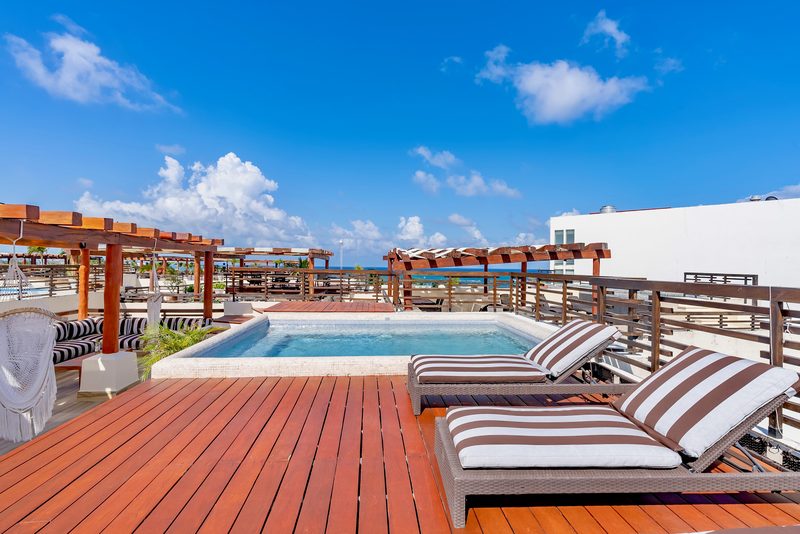 Plunge pool on a wood rooftop deck with two striped sun loungers in front, pergolas behind and a blue sky