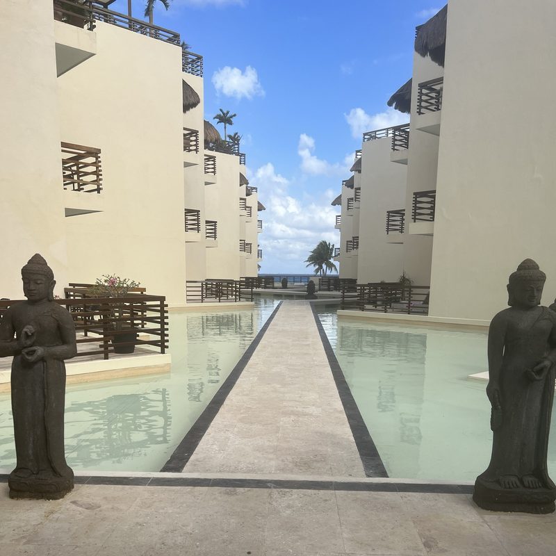 A stone walkway between reflecting pools and Buddha statues, leading to the sea, with white buildings either side